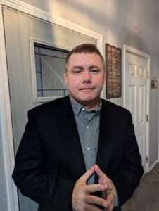 Professional headshot of a middle-aged man with short brown hair wearing a black suit jacket over a gray patterned dress shirt. He is standing in a hallway with white doors and trim, with his hands positioned in front of his chest. There is a decorative wall sign visible in the background.